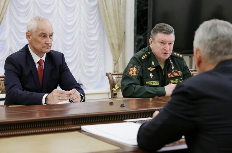 A man in a suit sits at a large wooden table near another man in a military uniform. 