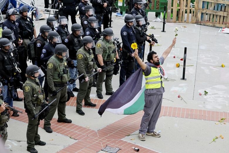 A man stands in front of a row of police officers. His arms are aloft and he hold flowers in both hands and wears a flag around his neck like a cape.