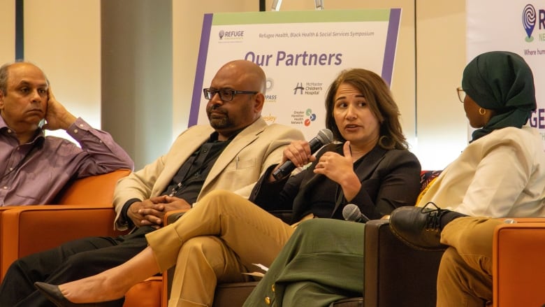 Four people sit in arm chairs facing forward while one speaks on a microphone. 