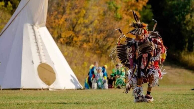 A man in traditional Indigenous regalia dancing with a teepee behind him. 