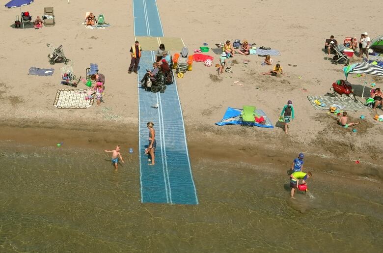 People are seen at a beach. Mats are on the sandy beach and go into the water's edge, helping people with wheelchairs cross the beach.