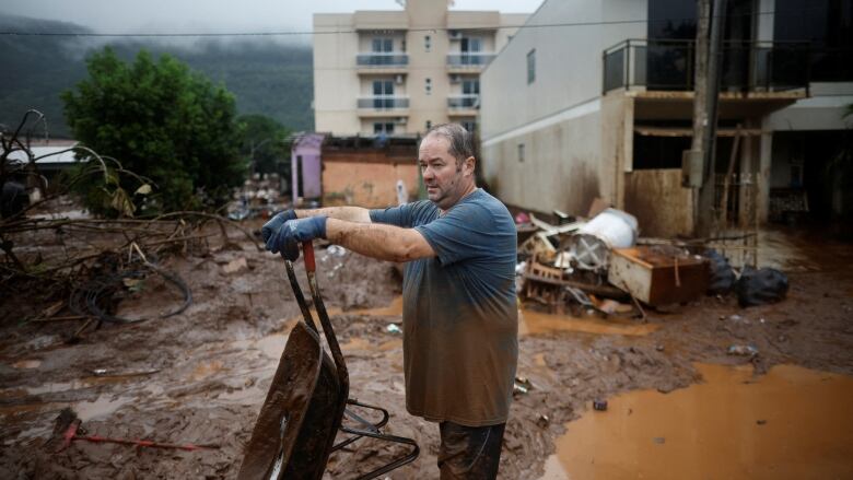 A man in muddy, wet clothing tips a wheelbarrow full of mud from inside his flooded home into a puddle. 