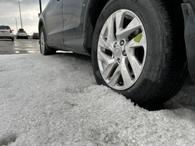 A car tire is seen close up next to a pile of hailstones in a parking lot.