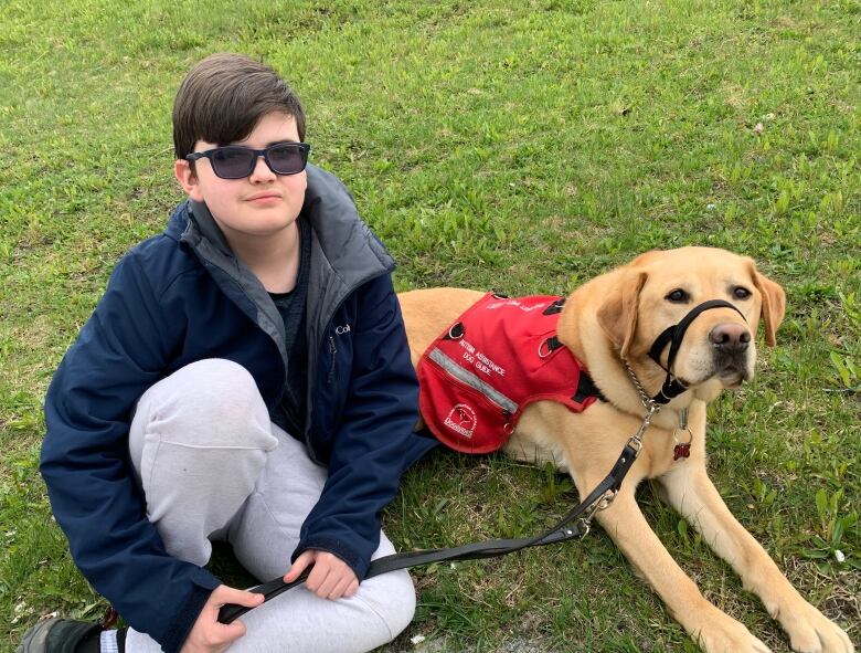 A pre-teen boy wearing grey sweatpants, a navy coat, and dark eyeglasses sits on the grass next to a golden retriever wearing a red vest. 