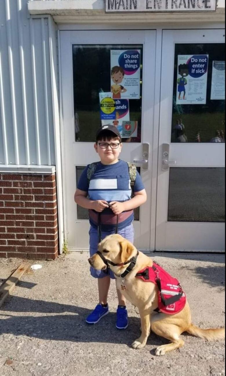 A boy of about 6 years old stands with a golden retriever outside the steel and glass doors of a school.