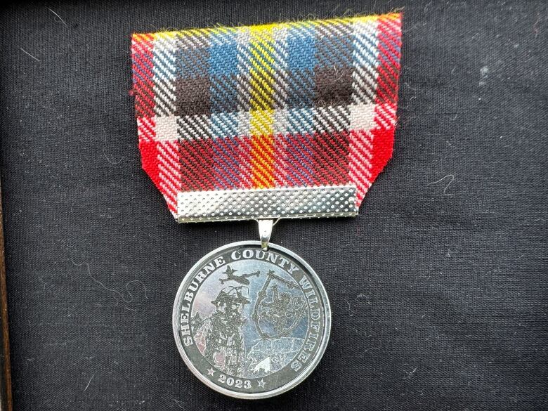 A silver medal is shown hanging from a tartan of the area of Barrington. A firefighter in a helmet and a plane are featured in the medal.