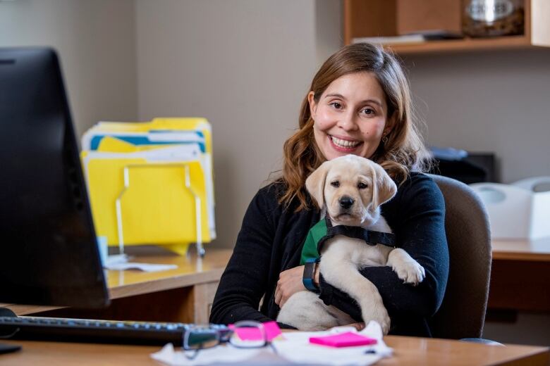 A smiling woman with long auburn hair sits at an office desk. She's holding a golden retriever puppy. 