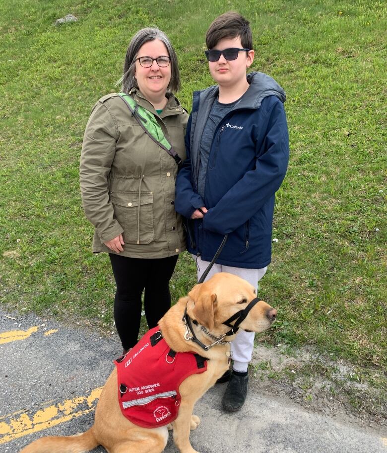 A woman wearing an olive green jacket stands next to a pre-teen boy wearing a navy blue coat. A golden retriever wearing a red vest sits on the ground in front of them.