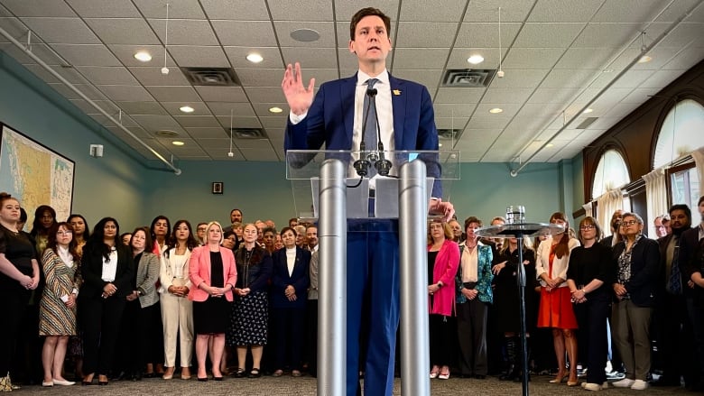 A man in a blue suit stand infront of a microphone with his hand raised. A crowd of people stand behind him.