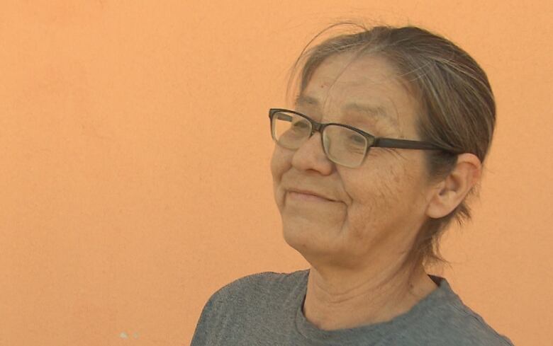 A woman in a grey T0shirt stands smiling in front of an orange wall outside.