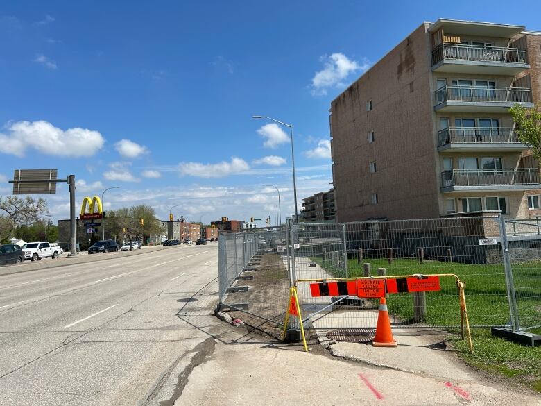 An orange barricade is shown in front of metal fencing surrounding a vacant apartment building.