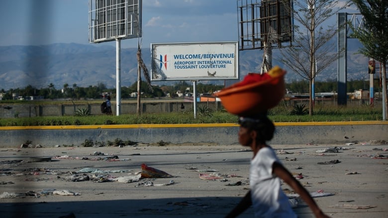 A blurred image of a woman on a street carrying a large, red bowl on her head as she walks past a welcome sign, in English and French, for an airport. Debris, garbage and clothing are scattered on the street below the sign.