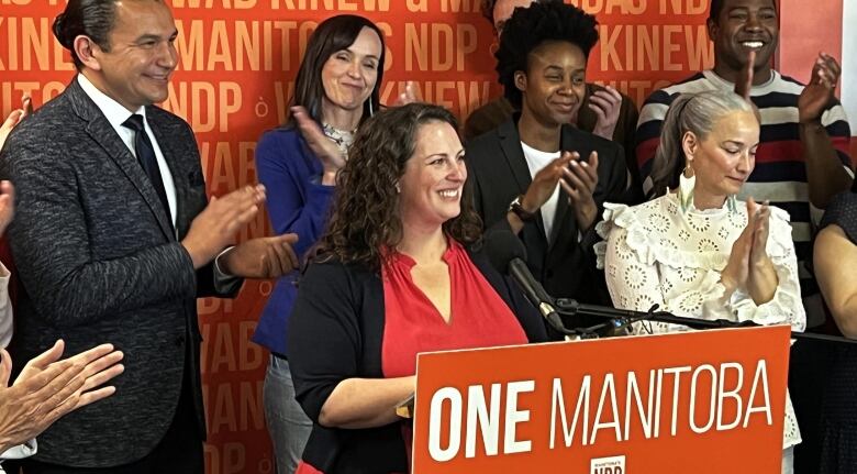 A woman standing at an orange podium smiles as she is surrounded by people clapping. 