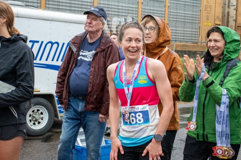 A person wearing a racing singlet and a medal around their neck is seen smiling in a crowd while it is raining.