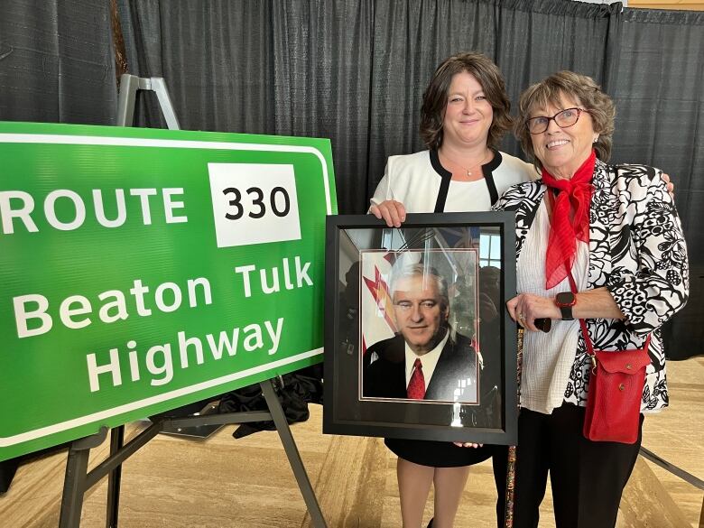 Two woman hold a picture of an older man next to a bright green sign that says Route 330 Beaton Tulk Highway.