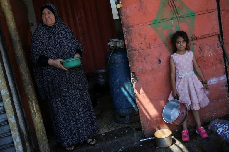 A woman and a young girl stand with empty bowls and pots in their hands.