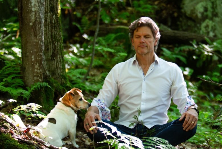 A man sits cross-legged in a forest clearing with his eyes closed. A small brown and white dog sits next to him.