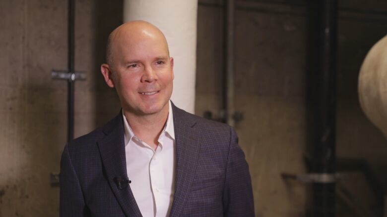 A bald white man smiles. He's wearing a suit jacket and white dress shirt with the top button unbuttoned. No tie.