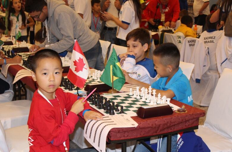 A young boy poses across the table with two others, playing chess, with flags of the world nearby.