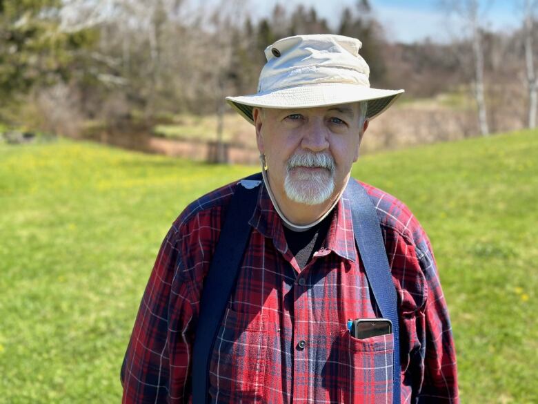 A man in a red plaid shirt and Tilly hat stands near a river. 