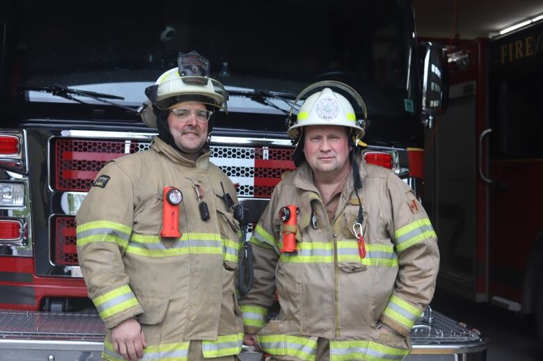 Two men in firefighting uniforms stand in front of a fire truck.