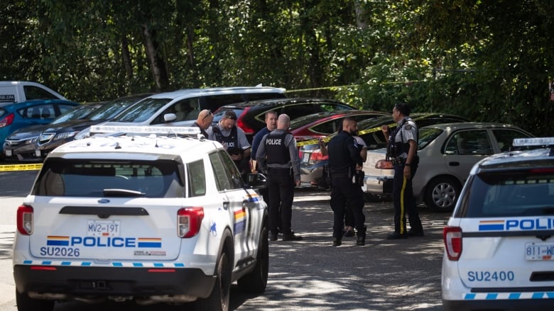 Two white police SUVs are seen from the rear in a parking lot with about eight police officers spread out in front of them having discussions and looking at notes.