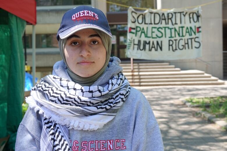 A woman wearing a ball cap and head covering starts at the camera with a serious face. A white and black keffiyeh scarf is around her shoulders. Tents and banners can be seen behind her.