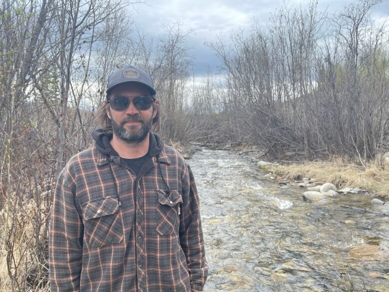 Troy Friday stands in front of Stony Creek. He's wearing a plaid overshirt, a baseball hat and sunglasses. In the background, the creek rushes through a small stand of trees.