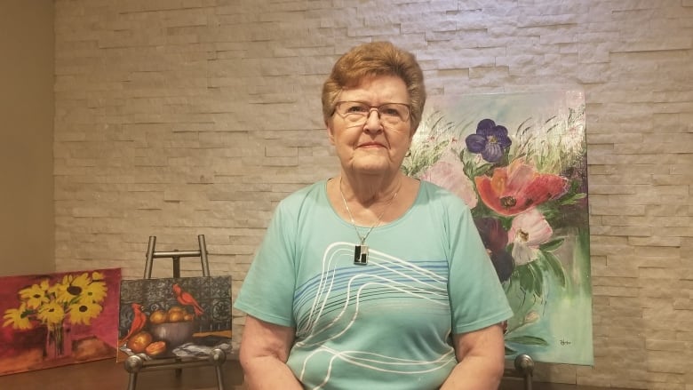 Shirly  is wearing glasses and a green shirt. She's looking directly at the camera and sitting in front of a stone wall and a display of paintings