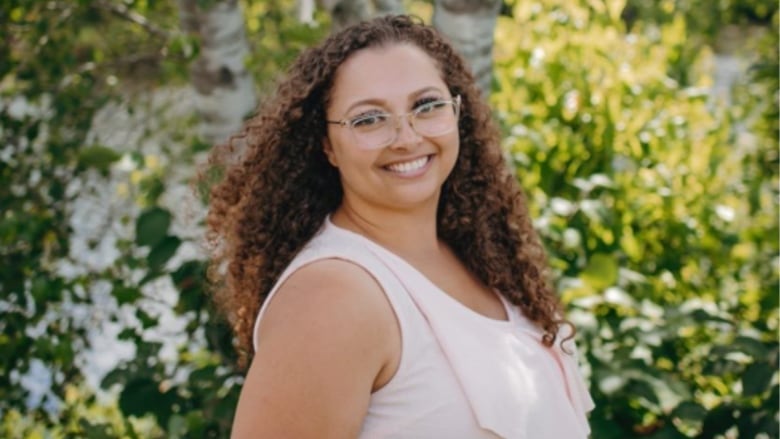 A woman with long brown curly hair wearing glasses and a tank top stands in a wooded area.