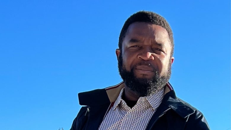 A man wearing a jacket and a white dress shirt stands near a blue background.