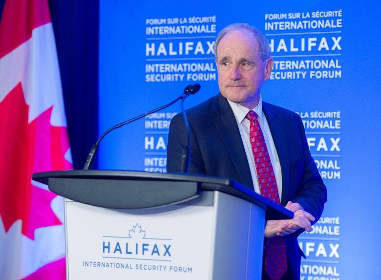 A man in a dark suit and red tie approaches a podium to speak.