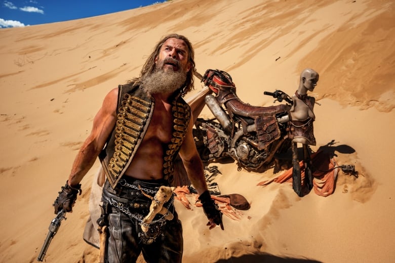 A shirtless man wearing a vest, leather pants and with a teddy bear attached to chains around his waist stands on a dune. He is holding a revolver. Behind him is a motorcycle. 