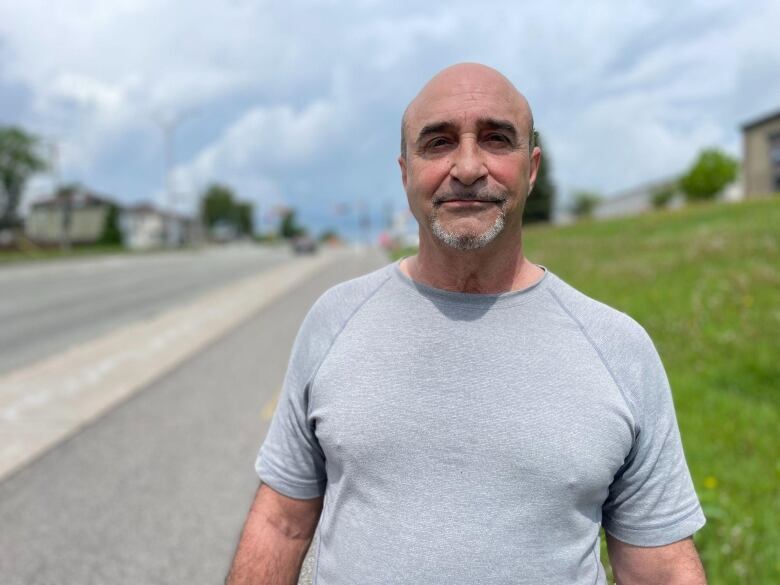 A man looks at the camera. He is standing on a sidewalk, outside.