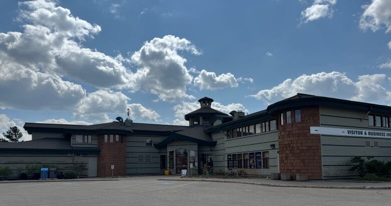 Grey one-story building with a small tower in the centre. The buildings two parts join each other at an obtuse angle. The pavement of the carpark in foreground. 