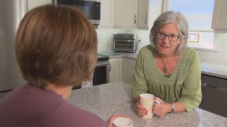 A woman with short light brown hair wearing a purple top sits at a kitchen counter with her back to the camera holding a white coffee cup. Across from her sits a woman with shoulder-length white hair, glasses and a green top, also holding a white coffee cup.