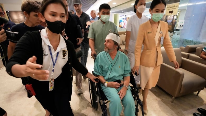 A man with a white bandage around his head sits in a wheelchair being pushed by medical staff.