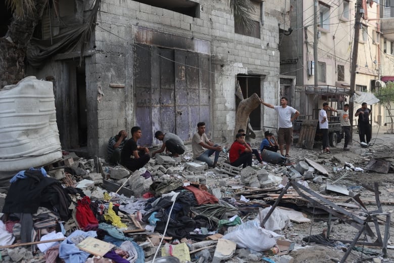 Several men sit against a wall near amid piles of concrete debris and strewn clothing. 