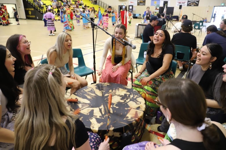 A group of girls seated around a traditional Indigenous drum, drumming and singing, with dancers in traditional Indigenous outfits in the background.