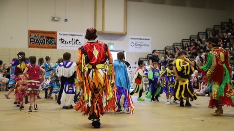 Sseveral students of various ages dressed in traditional Indigenous outfits dance in a school gym.