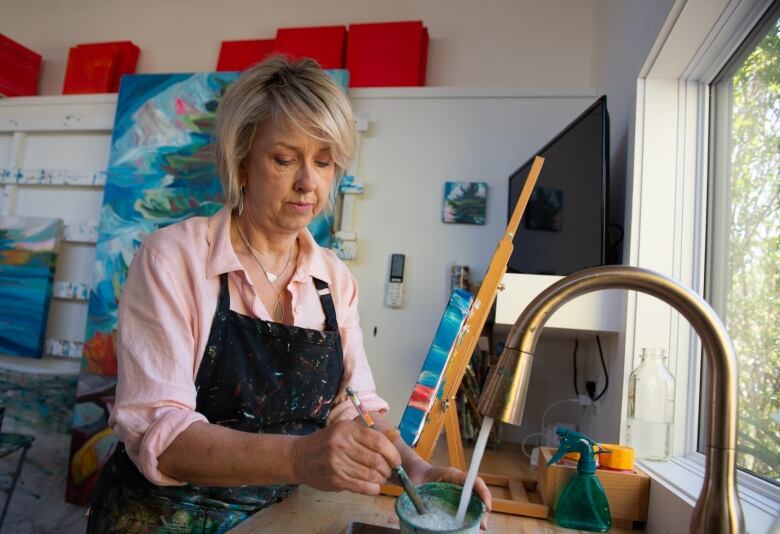 Woman in a pink shirt washes her paint brush in a sink in a brightly lit studio