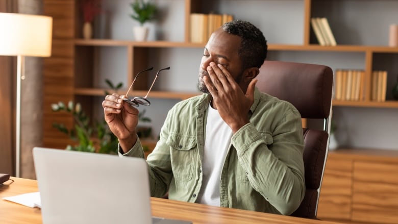 A man sitting at a desk in front of a laptop is rubbing his eyes while holding his glasses in one hand.