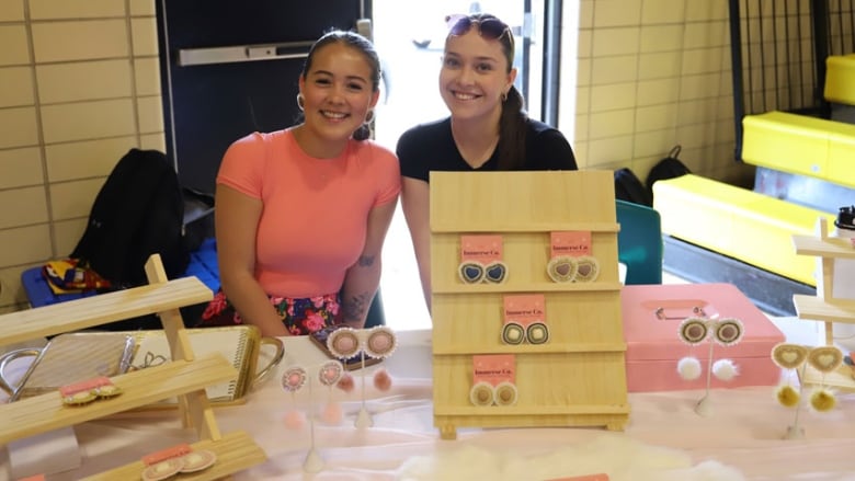 Two smiling teenaged girls seated at a table with Indigenous jewerly on display.