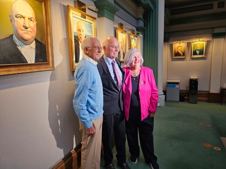 Three people stand together with their arms around each other inside the House of Assembly. They pose in front of a photo hanging on the wall.