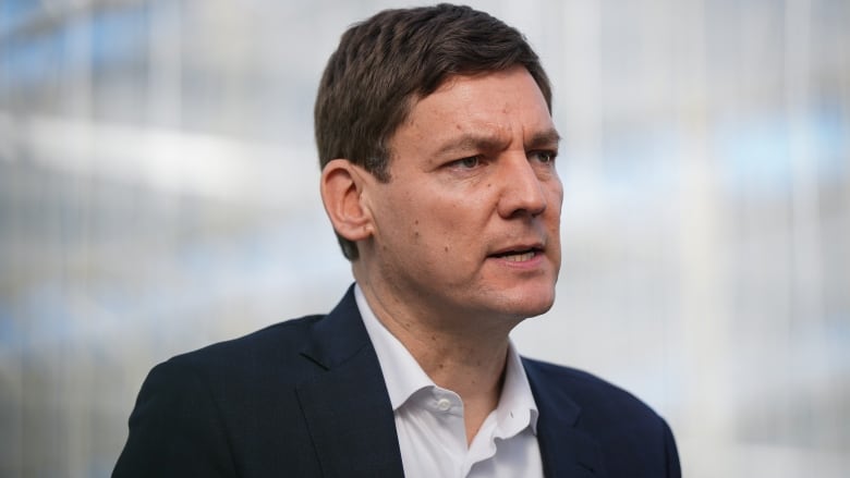 A portrait of B.C. Premier David Eby, a tall white man wearing a suit, speaking during an announcement