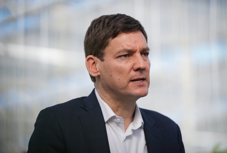 A portrait of B.C. Premier David Eby, a tall white man wearing a suit, speaking during an announcement