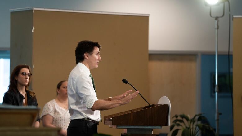 A man in white shirt and green tie speaks into a microphone at a podium. Two women look on.