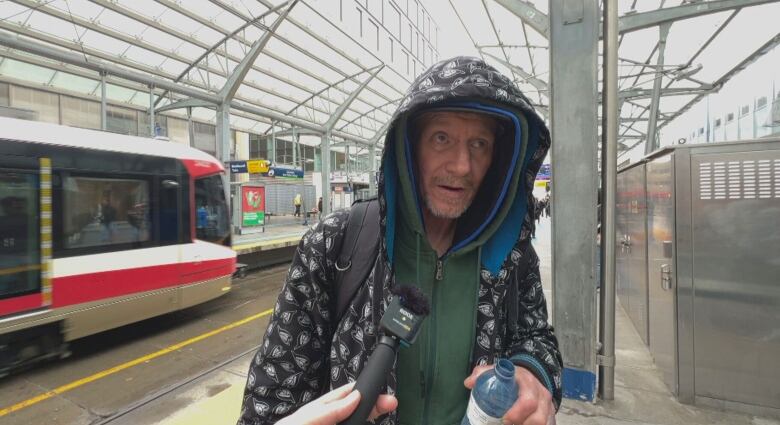 A man holding a water bottle stands on a C-train platform. He has a hood on to protect from the rain. 