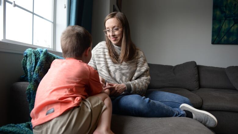 A woman with long, brown hair and glasses smiles as she sits on a sofa playing with a young boy whose back is to the camera. 