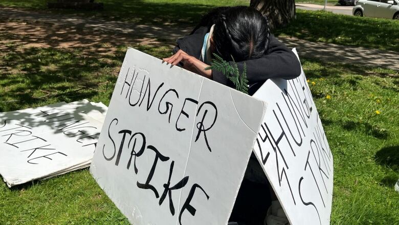 A protesters, head bowed, sits in the grass with hunger strike signs.